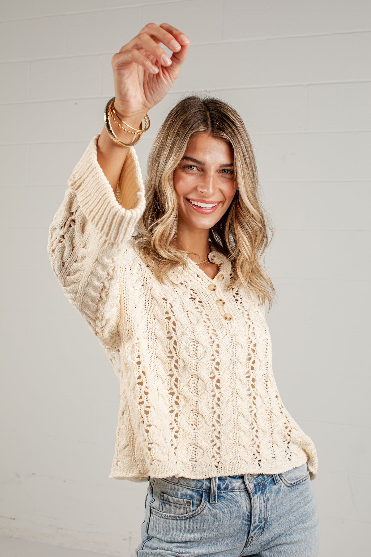A woman wearing a cream cable-knit button-up sweater and light-wash blue jeans, posing with one arm raised against a white paneled wall.