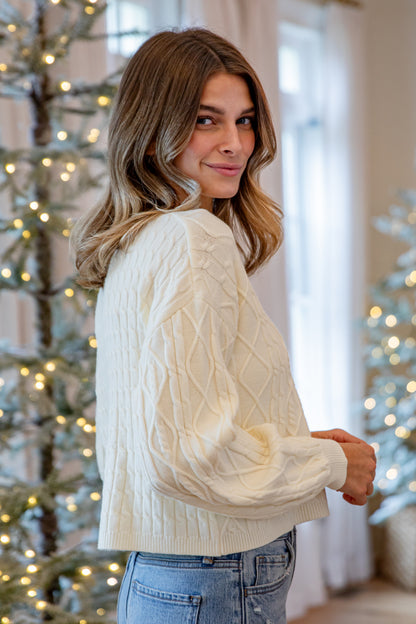 Woman wearing a white cable knit sweater in a festive indoor setting with Christmas trees.