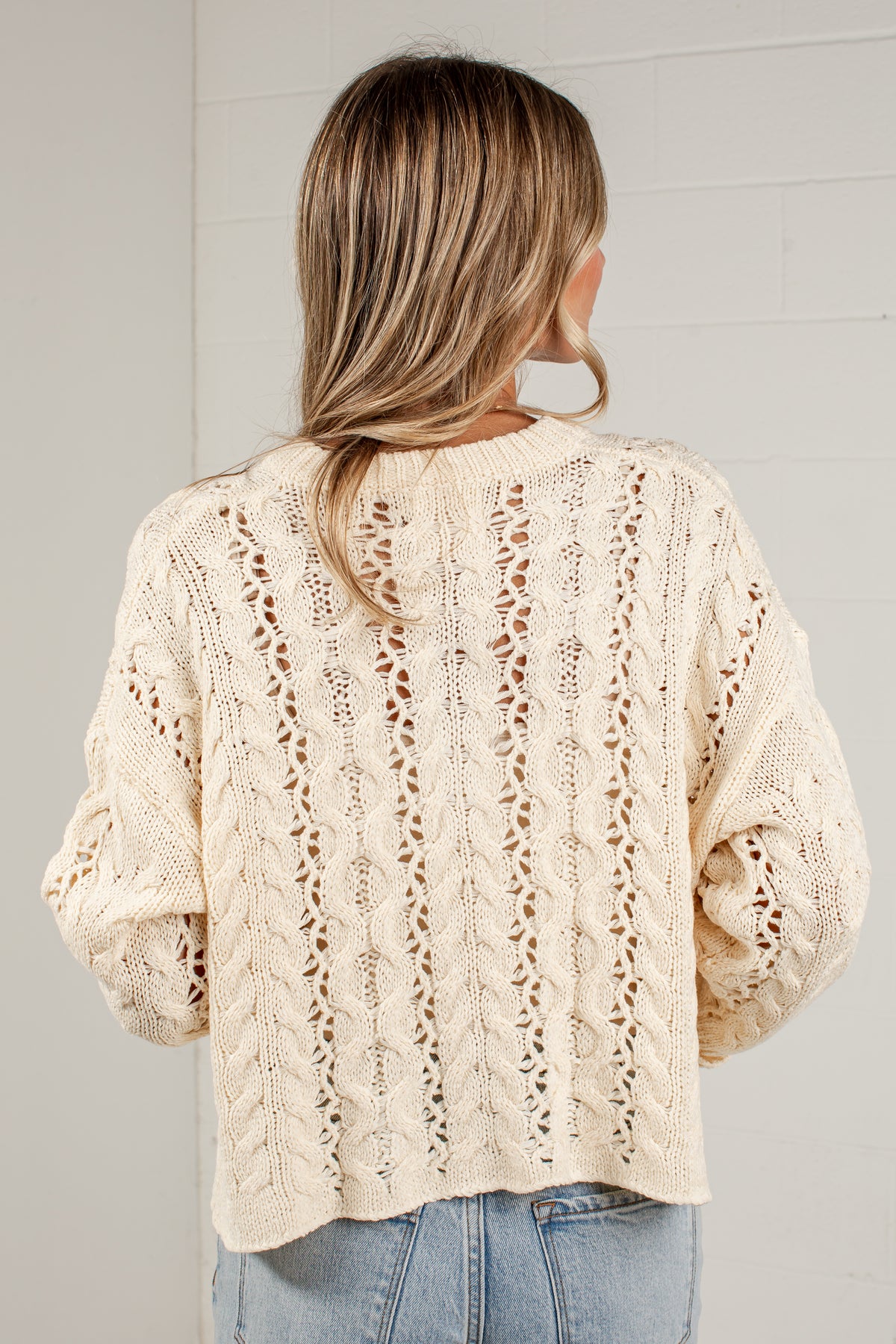 A woman viewed from behind wearing a cream-colored, cable-knit cropped sweater with textured openwork patterns, paired with light blue jeans, against a light gray brick wall.