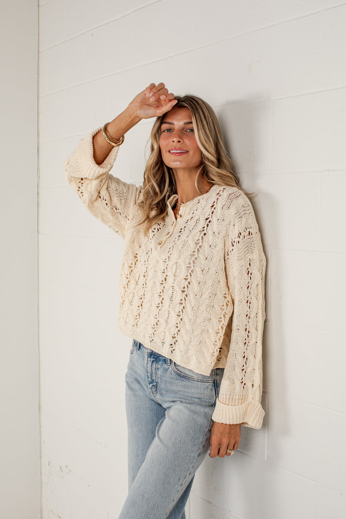 A woman wearing a cream cable-knit long-sleeve sweater and light-wash blue jeans, leaning against a white paneled wall.