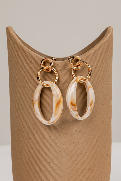 Pair of gold and marbled beige hoop earrings on a brown stand.
