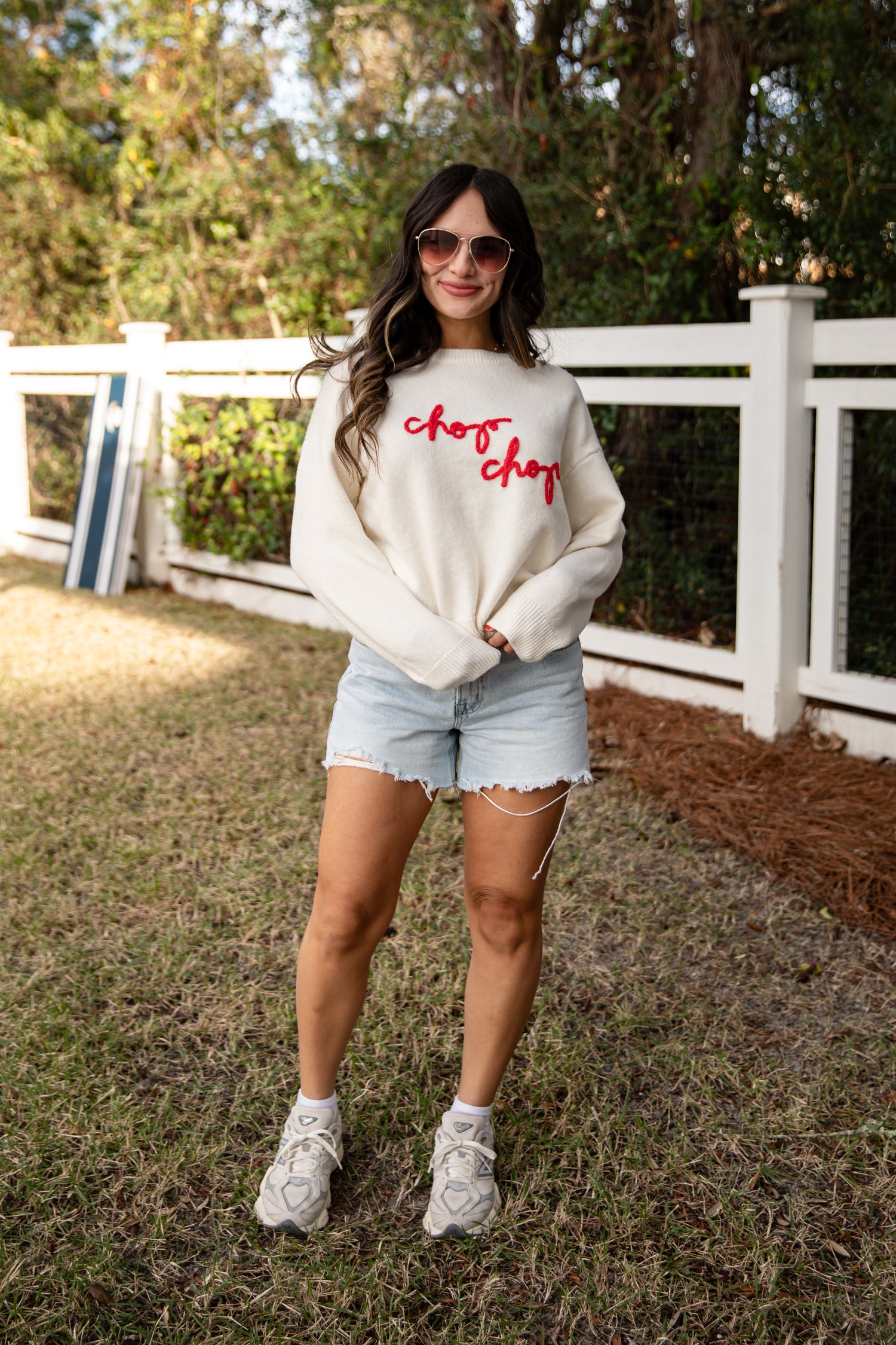 Woman wearing a white sweatshirt with red text and denim shorts outdoors.