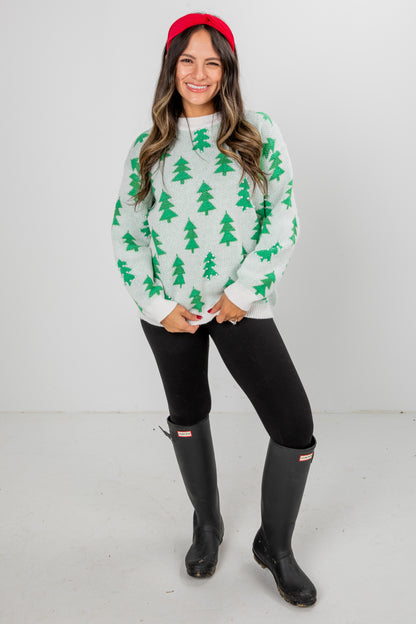 Woman wearing a white sweater with green tree patterns, black leggings, and black boots on a white background