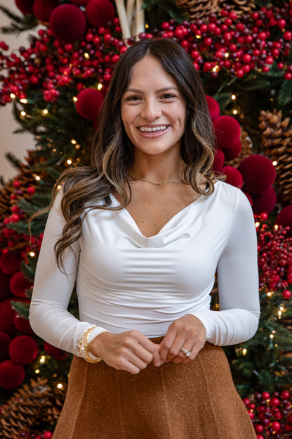 Woman in a white long-sleeve top and brown skirt standing in front of a festive background with red berries and lights.