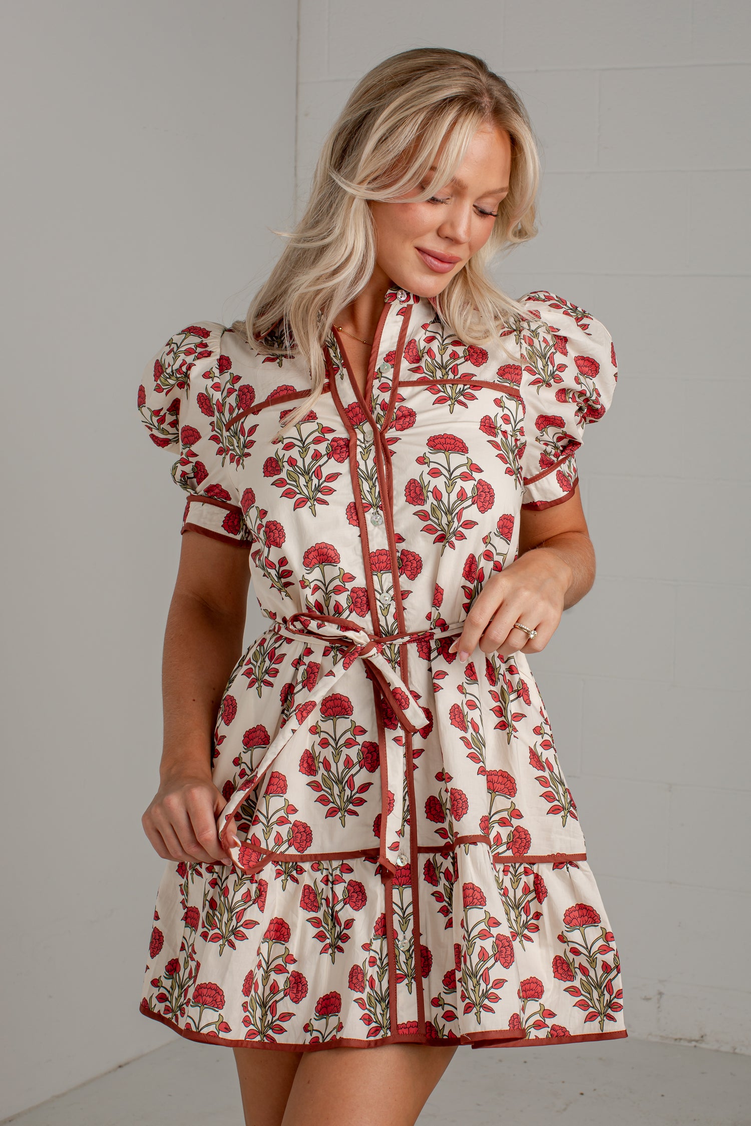 A woman modeling a short cream dress with a red and green floral print, featuring puff sleeves, a tie-neck collar, and a matching waist tie, set against a light gray wall.