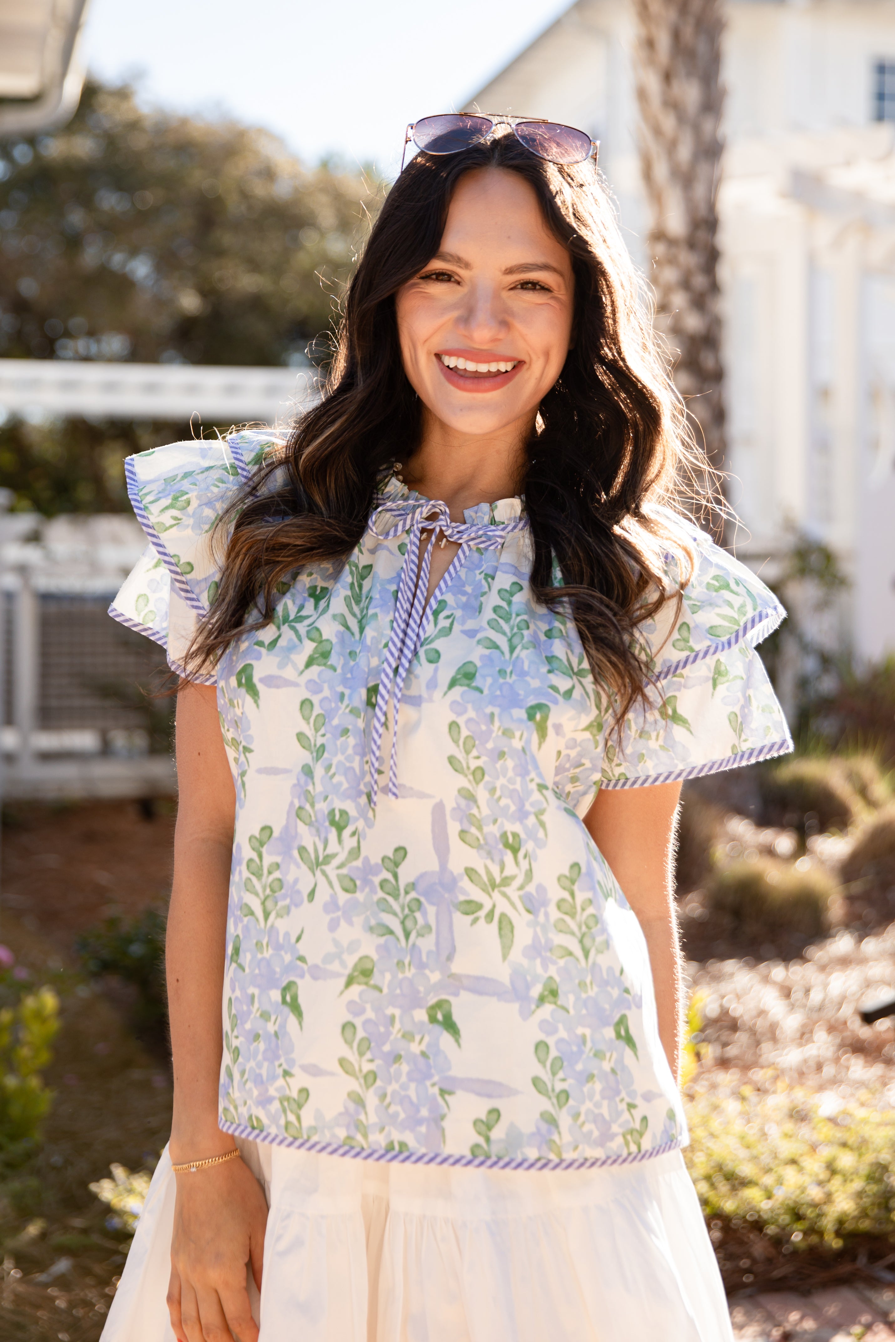 Woman wearing a floral blouse outdoors with greenery and a house in the background