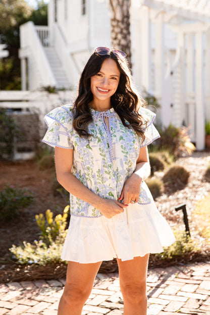 Woman in a floral top and white skirt standing outdoors with a white house in the background