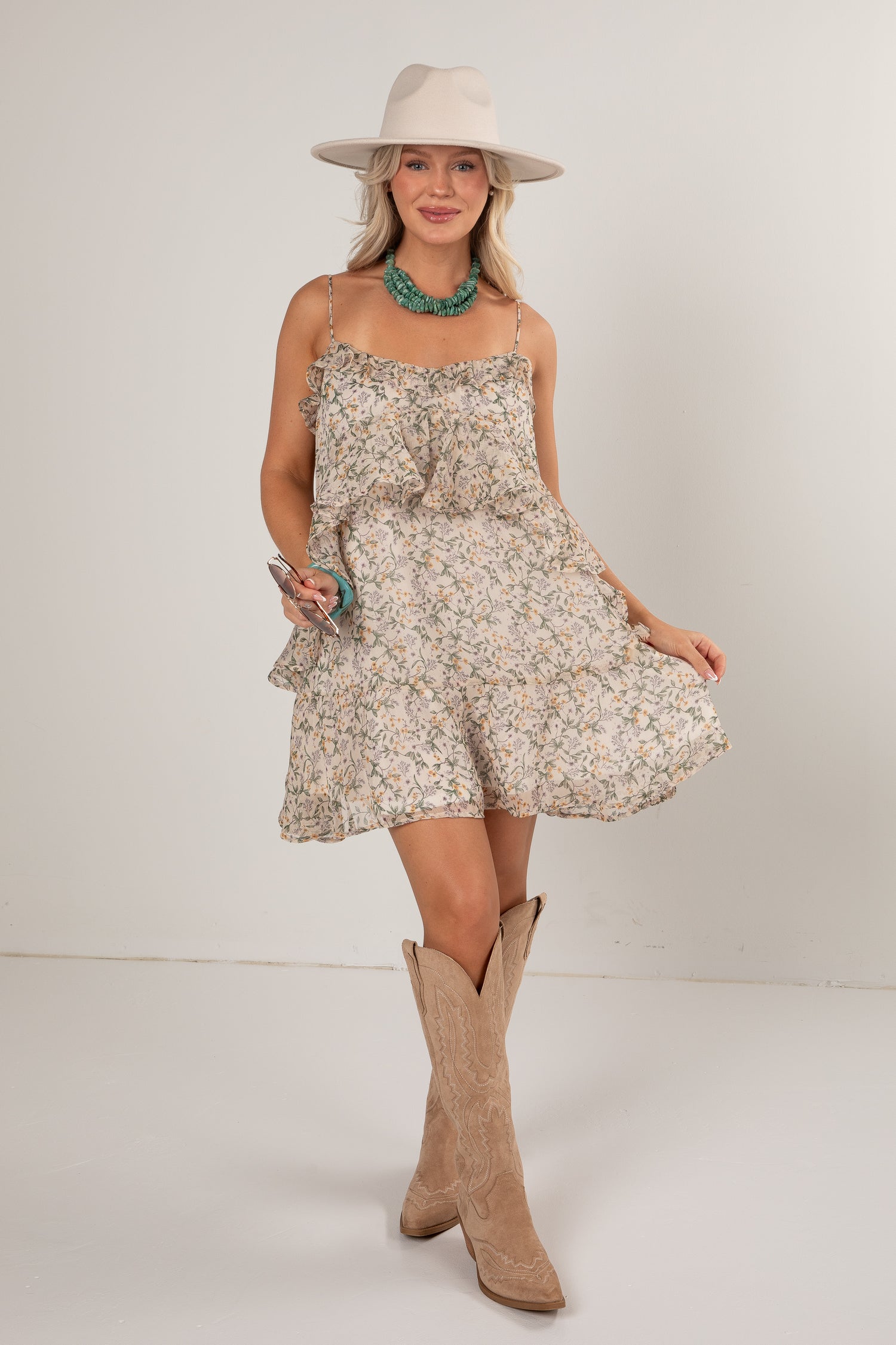 A woman modeling a short sleeveless ruffle-trimmed dress with a beige and white floral print, styled with a cream wide-brim hat, turquoise necklace, and tan cowboy boots against a plain light gray studio background.