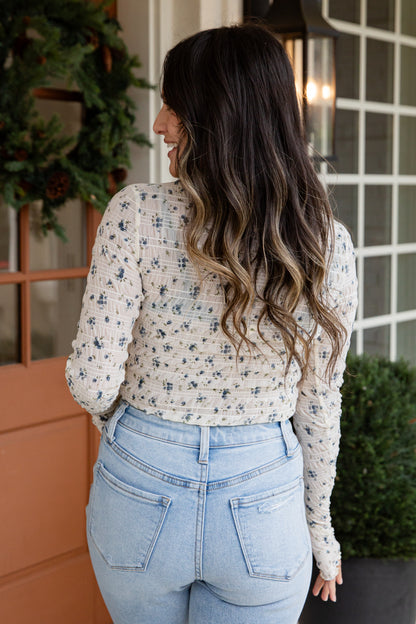 Woman wearing a floral long-sleeve top and light blue jeans, standing in front of a building with decorative elements.