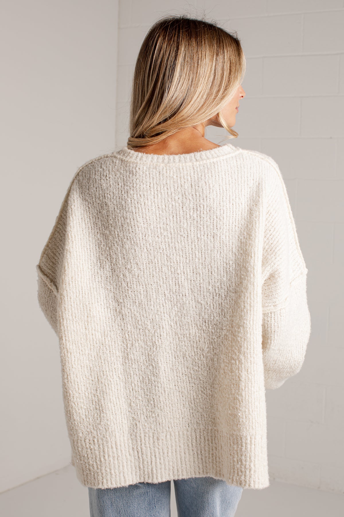 A woman viewed from behind wearing an oversized, textured cream knit sweater and light blue jeans, standing against a white brick wall.
