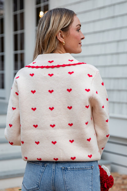 Woman wearing a cream sweater with red heart patterns and blue jeans.