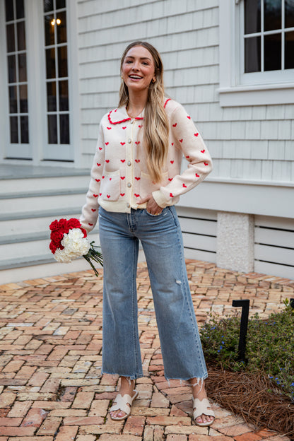 Woman wearing a heart-patterned cardigan and jeans, standing on a brick path in front of a house.