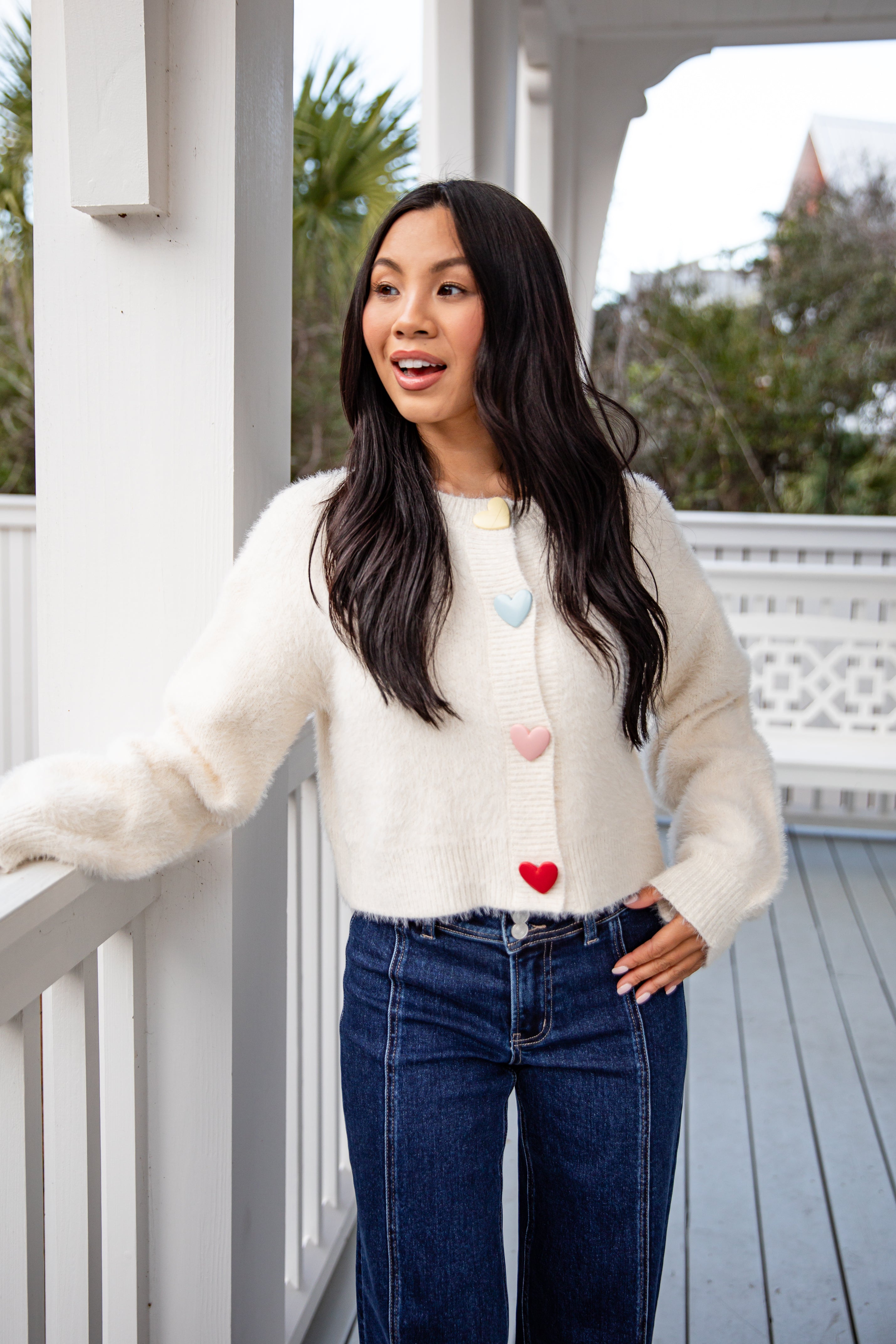 Woman wearing a white sweater with colorful buttons and blue jeans on a porch.