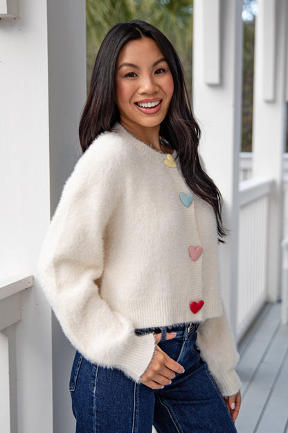 Woman wearing a cream sweater with heart patterns, standing on a wooden deck.