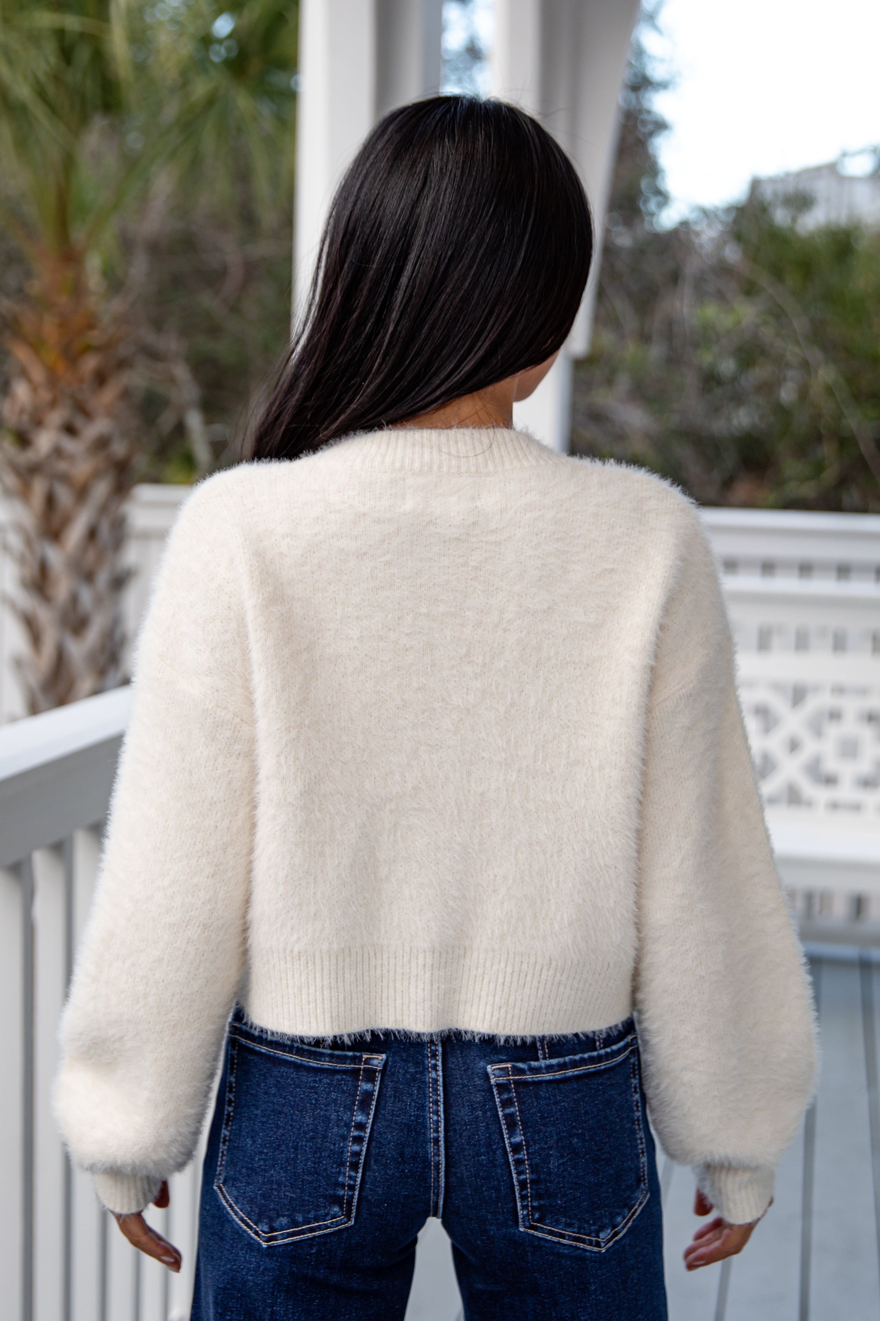 Person wearing a cream sweater and blue jeans standing on a porch.