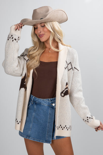 Woman wearing a patterned cardigan, brown top, and denim skirt with a cowboy hat on a gray background