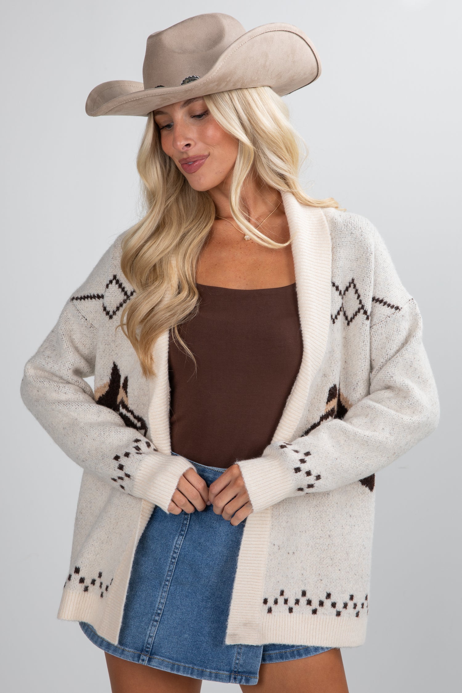 Woman wearing a beige cardigan with star patterns, brown top, and denim skirt on a gray background