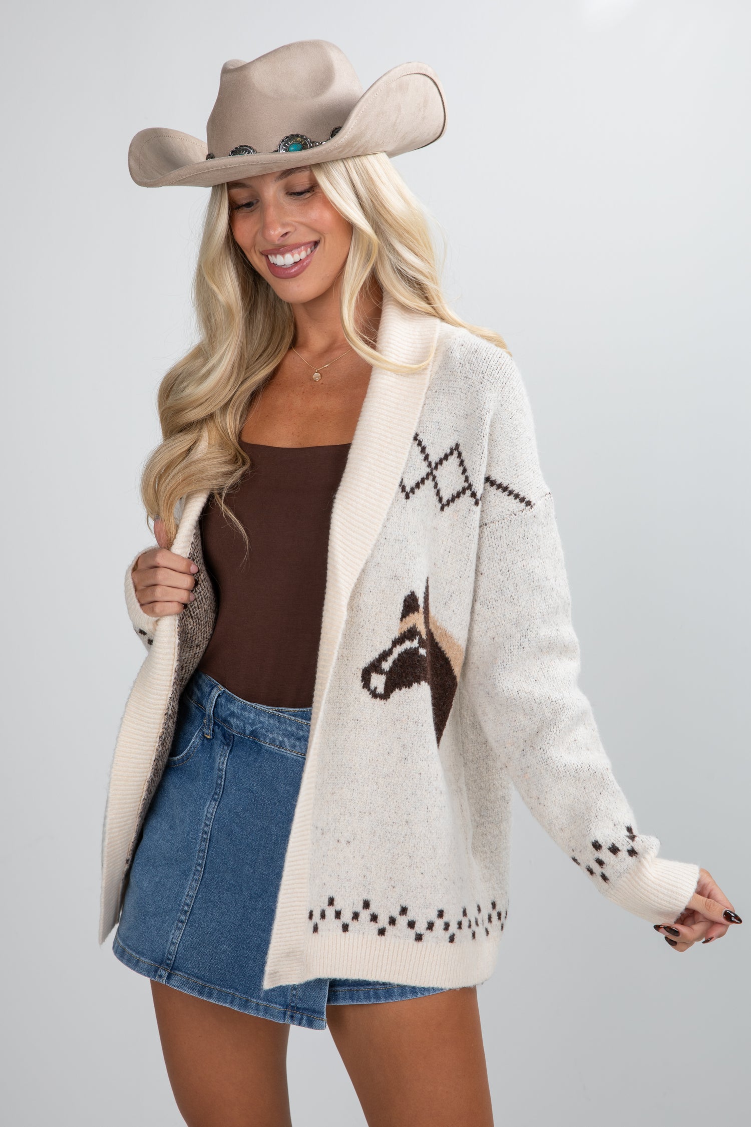 Woman wearing a patterned cardigan, denim skirt, and cowboy hat on a plain background