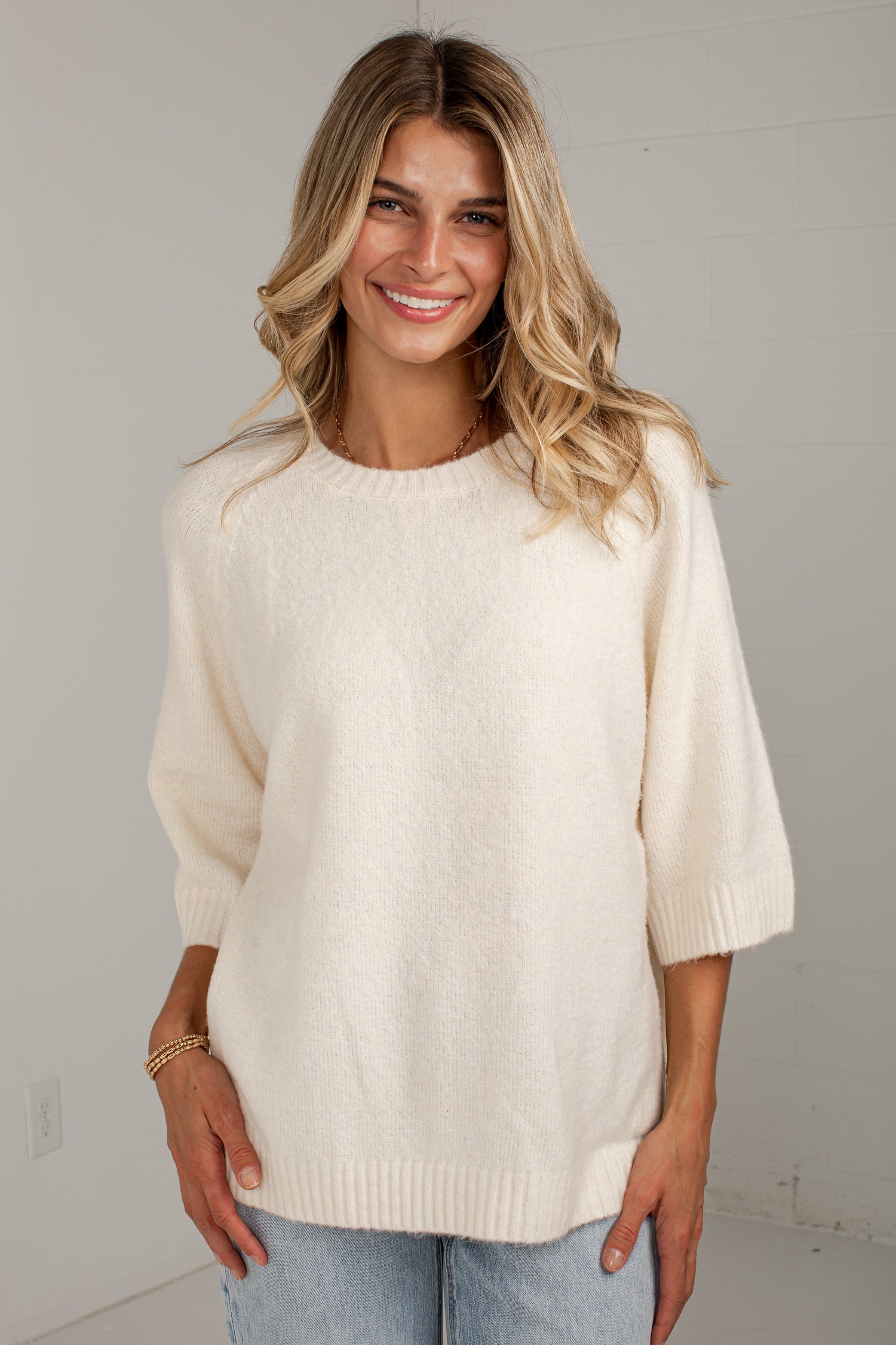 A woman wearing a cream-colored, fuzzy knit sweater with three-quarter sleeves and a crew neck, paired with light blue jeans, standing against a plain light gray wall.