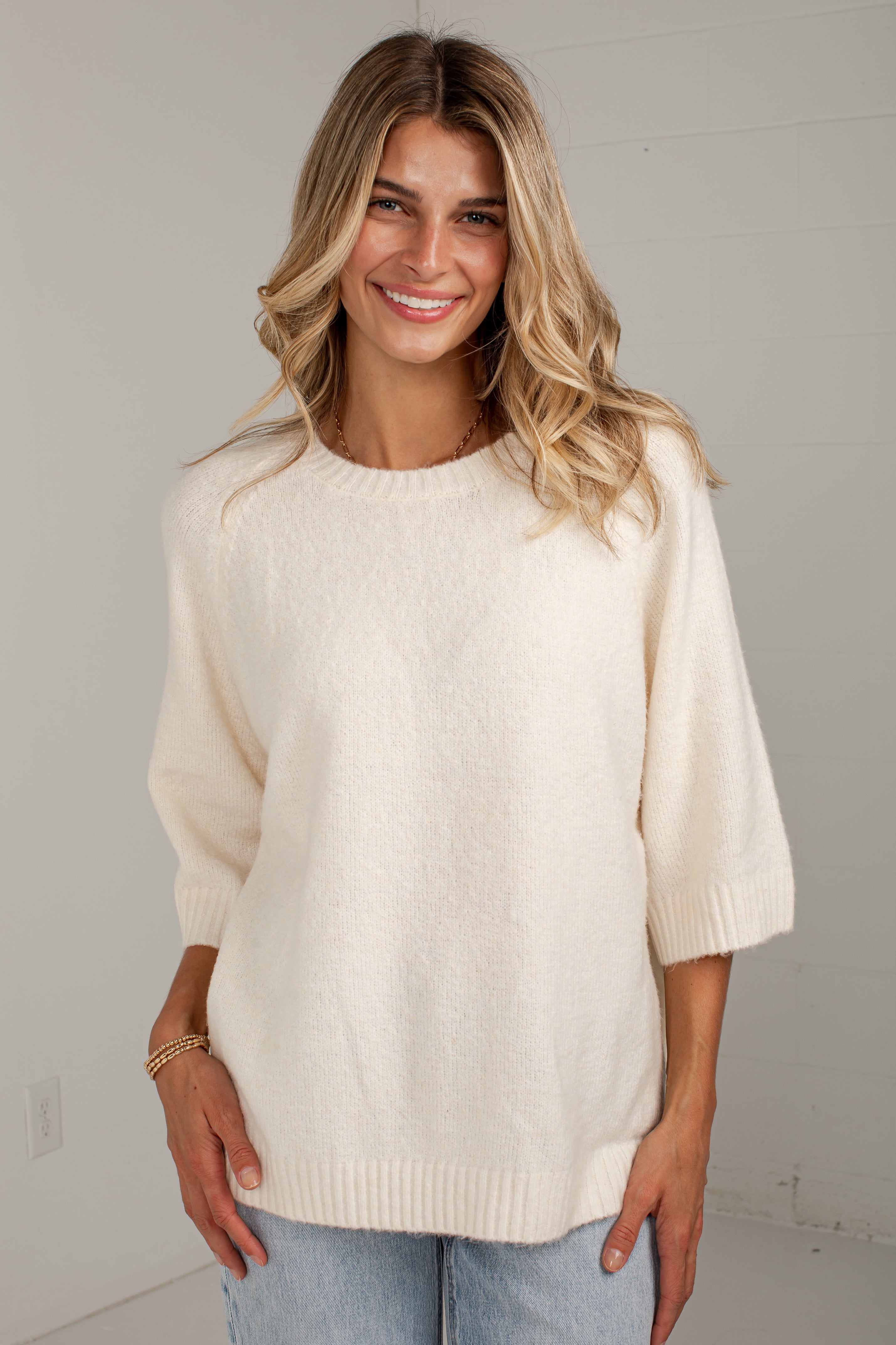 A woman wearing a cream-colored, fuzzy knit sweater with three-quarter sleeves and a crew neck, paired with light blue jeans, standing against a plain light gray wall.