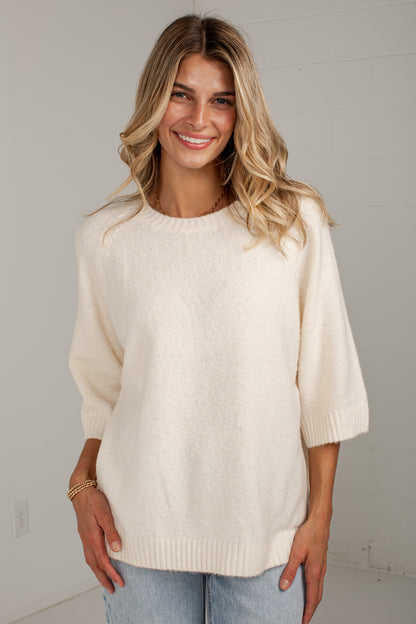A woman wearing a cream-colored, fuzzy knit sweater with three-quarter sleeves and a crew neck, paired with light blue jeans, standing against a plain light gray wall.