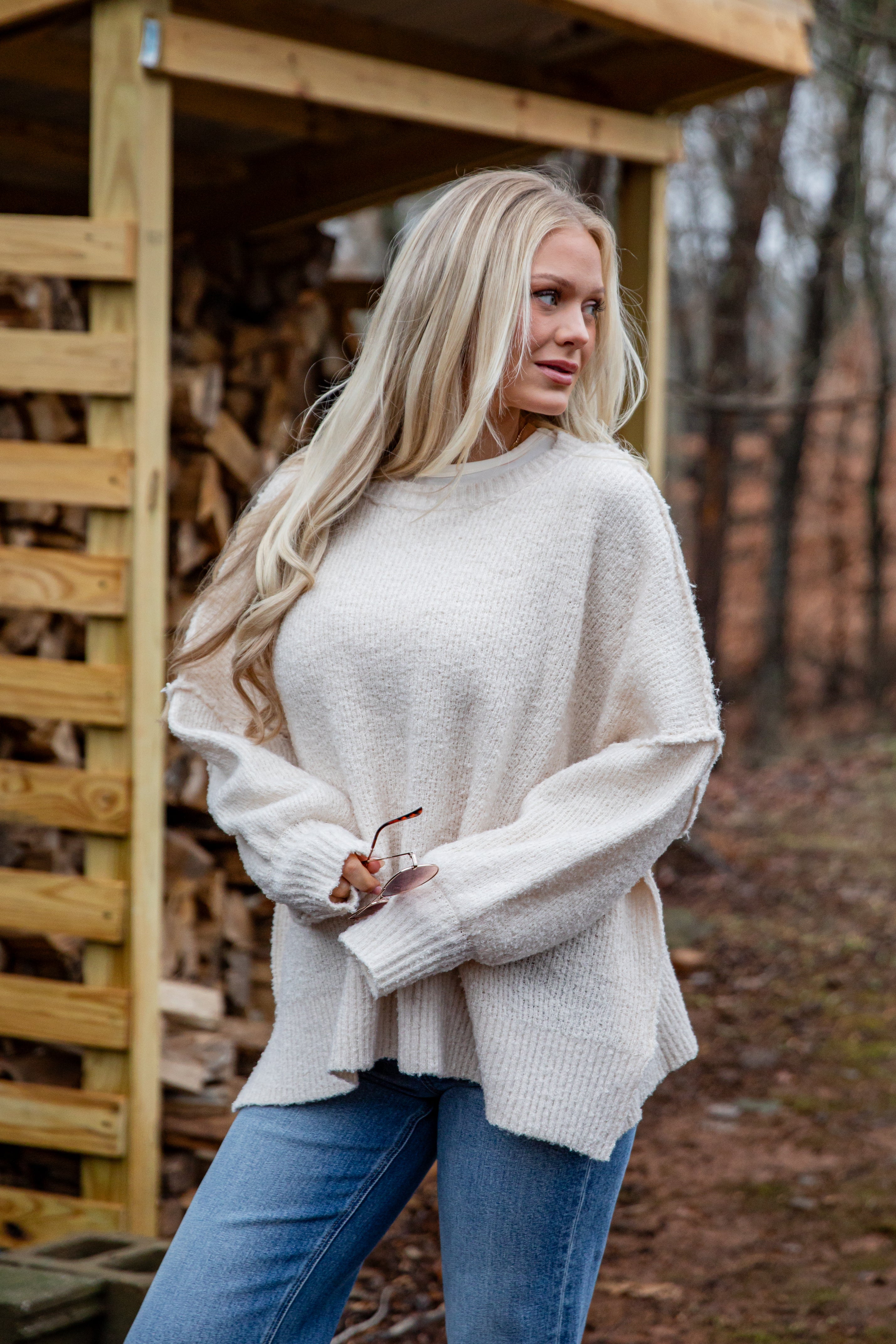 Woman wearing a white sweater and blue jeans standing in front of a wooden structure with stacked firewood.