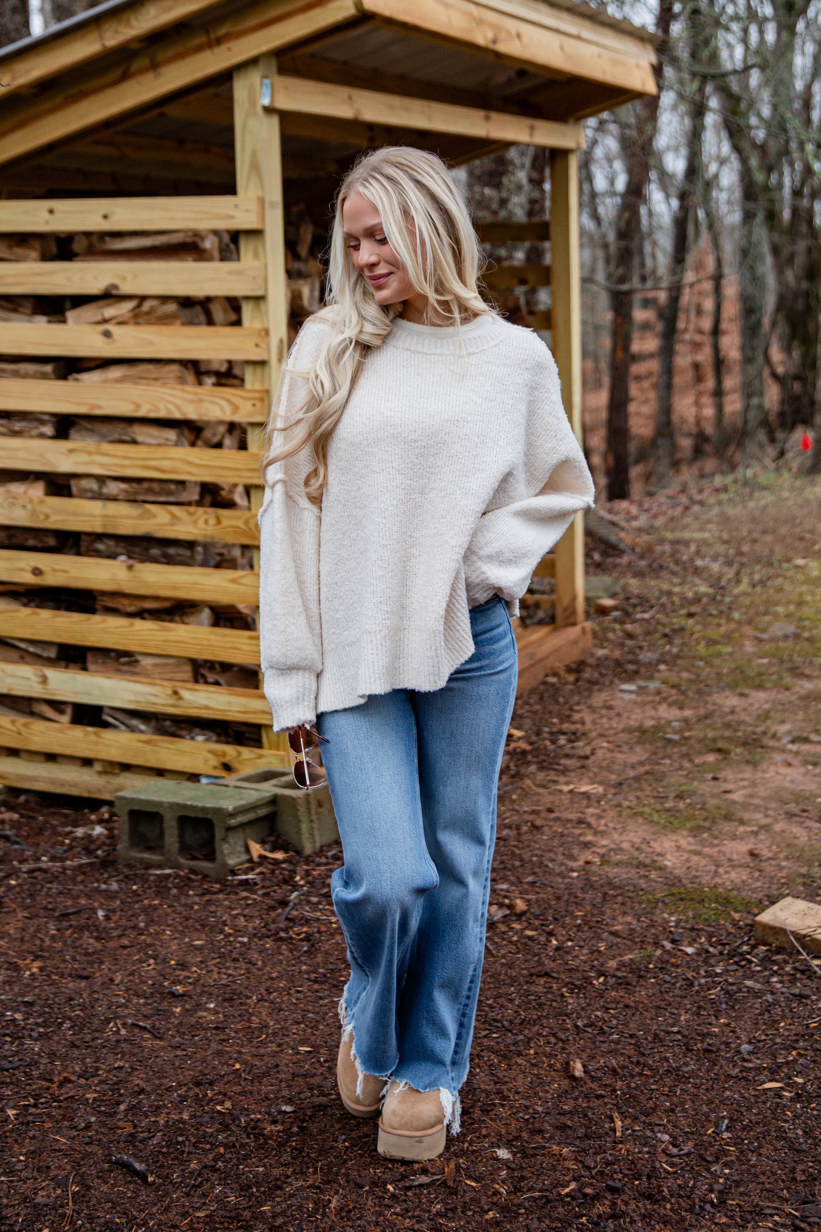 Woman in a white sweater and blue jeans standing in front of a wooden shed.