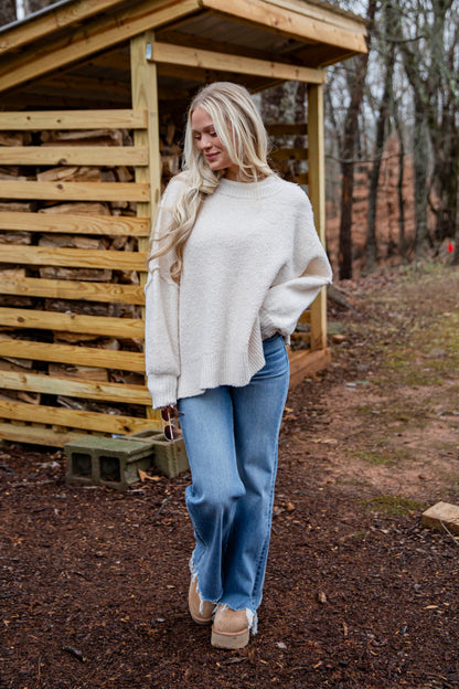 Woman in a white sweater and blue jeans standing in front of a wooden shed.