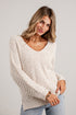 A woman wearing a chunky knit cream-colored long-sleeve sweater and light blue jeans, standing against a white paneled wall.