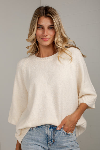 A woman wearing a cream-colored, oversized knit sweater and light blue jeans, standing against a light gray wall.
