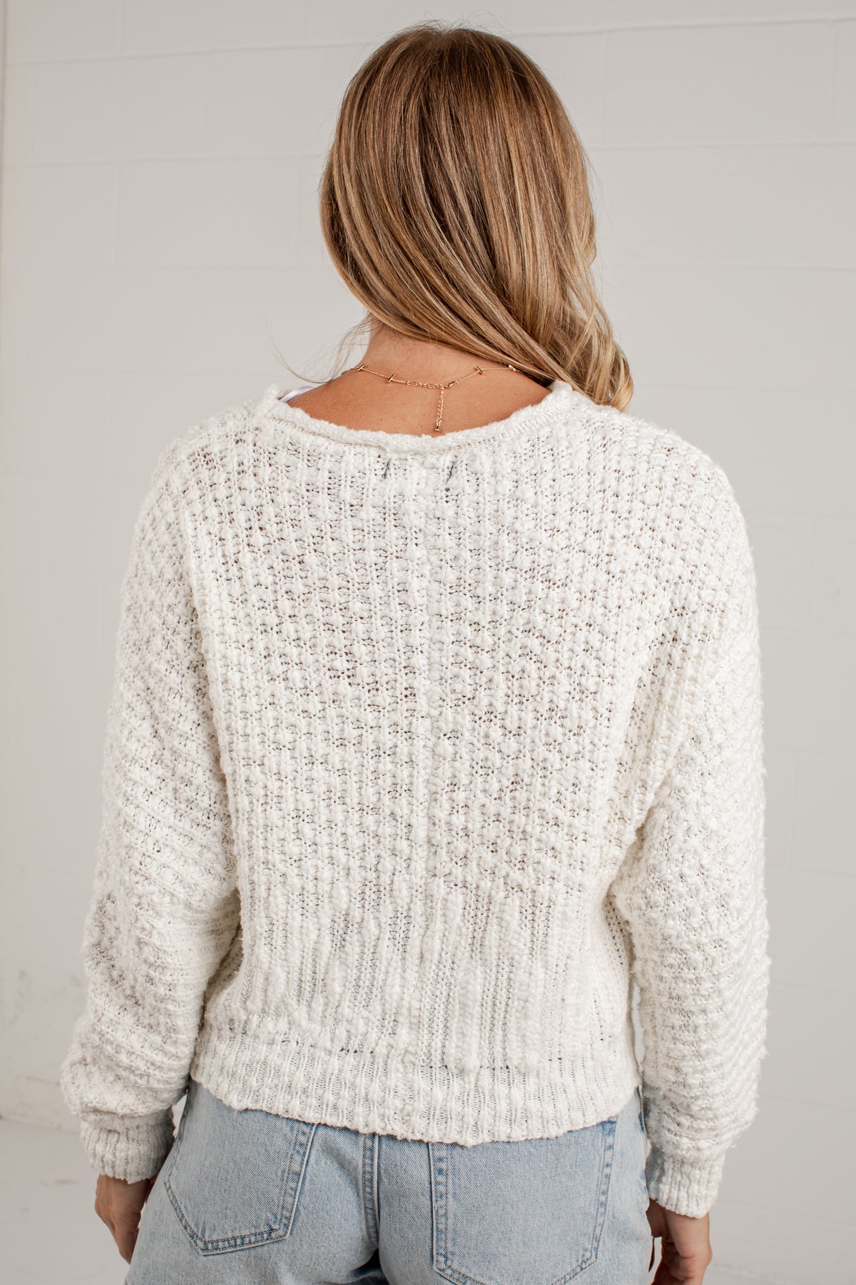 A woman viewed from behind wearing a chunky cream-colored cable-knit sweater and light-wash blue jeans against a white paneled wall.