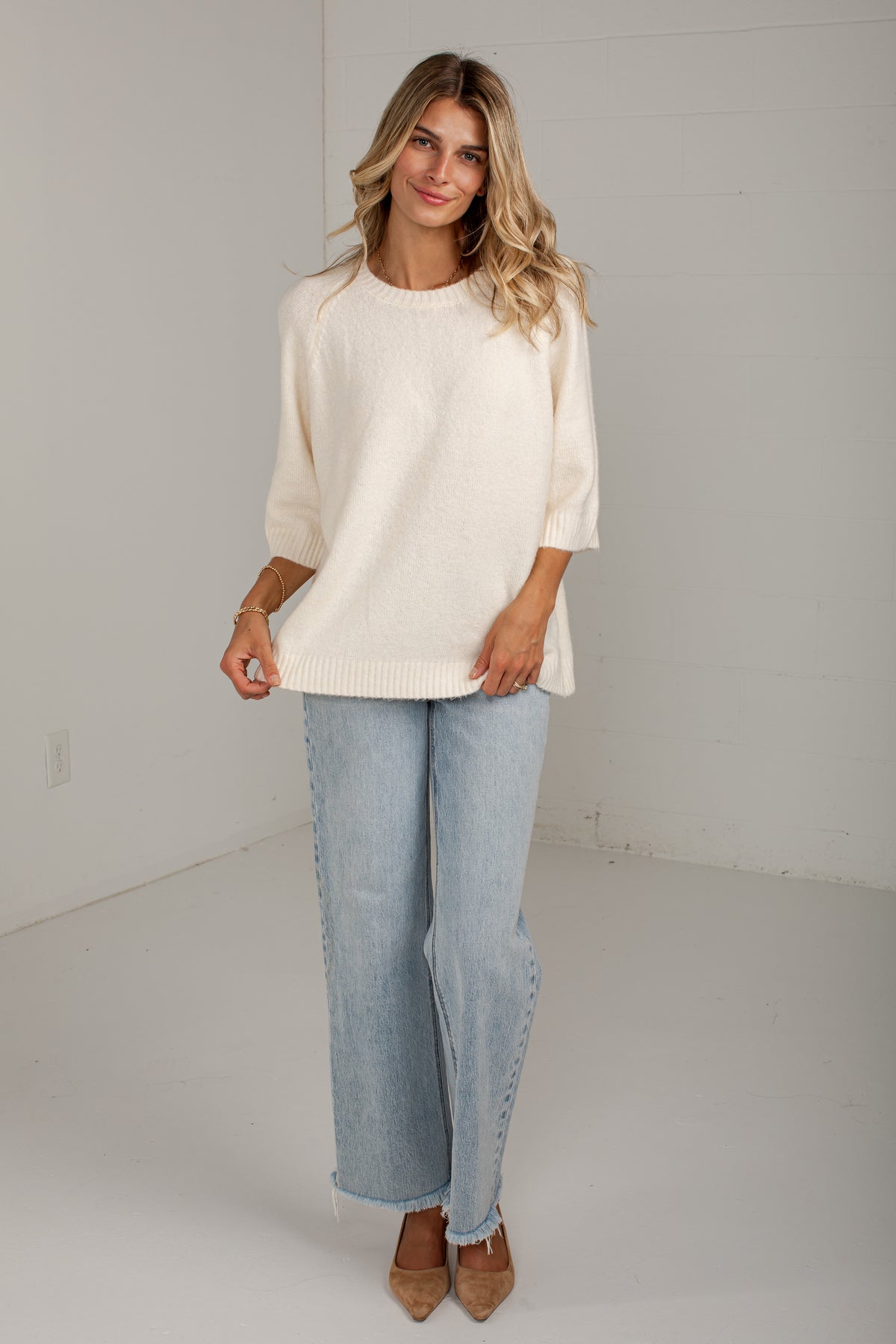 A woman modeling a cream-colored knit sweater with three-quarter sleeves, paired with light-wash wide-leg jeans and tan pointed-toe heels, standing against a white studio wall.