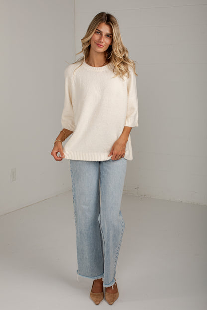 A woman modeling a cream-colored knit sweater with three-quarter sleeves, paired with light-wash wide-leg jeans and tan pointed-toe heels, standing against a white studio wall.