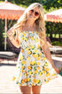 Woman in a lemon print dress standing outdoors with umbrellas and greenery in the background