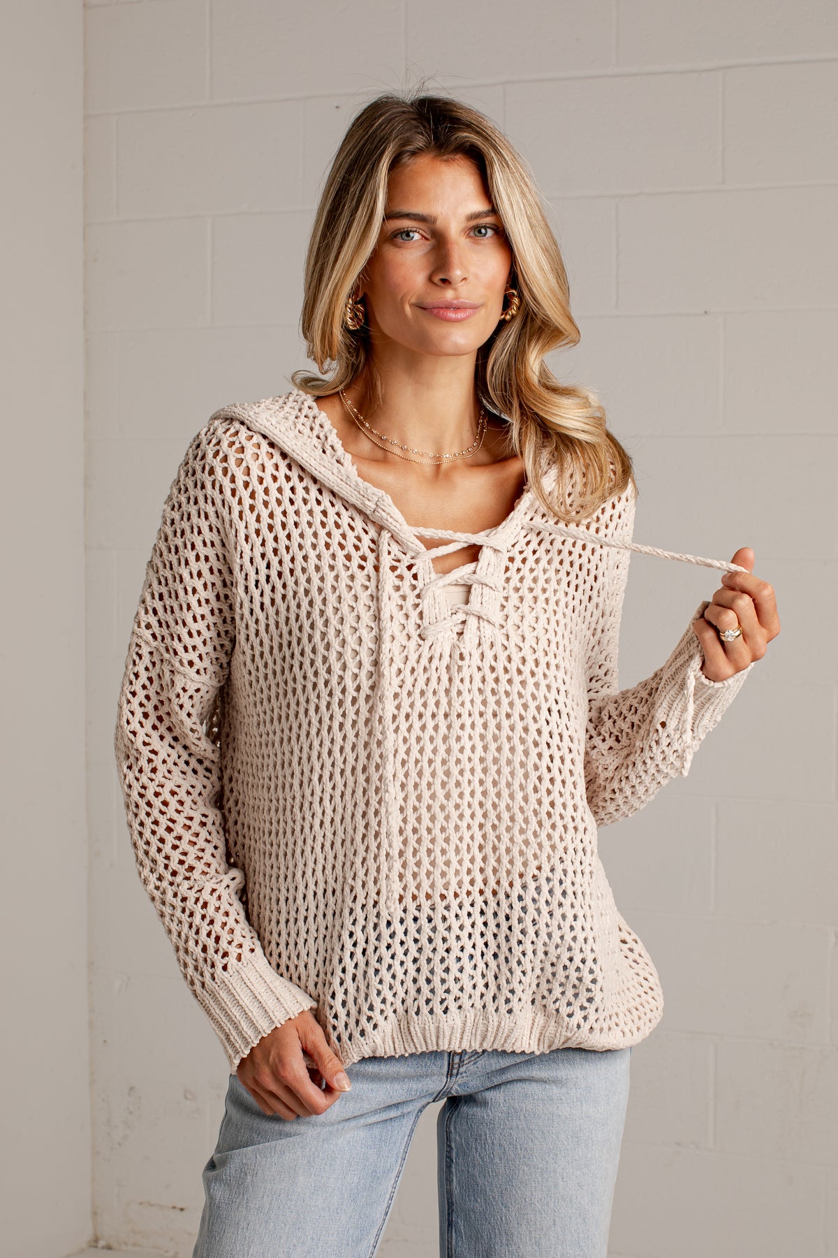 A woman wearing a cream-colored, open-knit crochet hooded sweater with lace-up front details and drawstrings, paired with light-wash blue jeans, standing against a light gray brick wall.