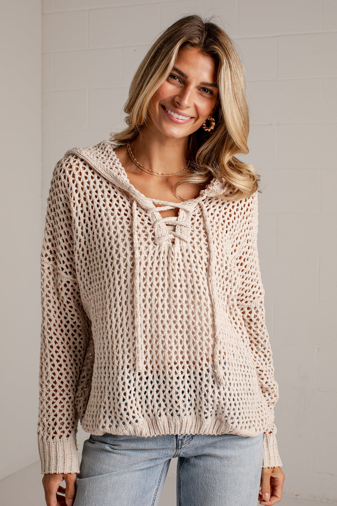 A woman wearing a cream-colored, open-knit hooded sweater with lace-up front details, paired with light-wash blue jeans, standing against a light gray brick wall.