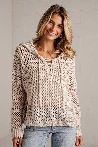 A woman wearing a cream-colored, open-knit hooded sweater with lace-up front details, paired with light-wash blue jeans, standing against a light gray brick wall.