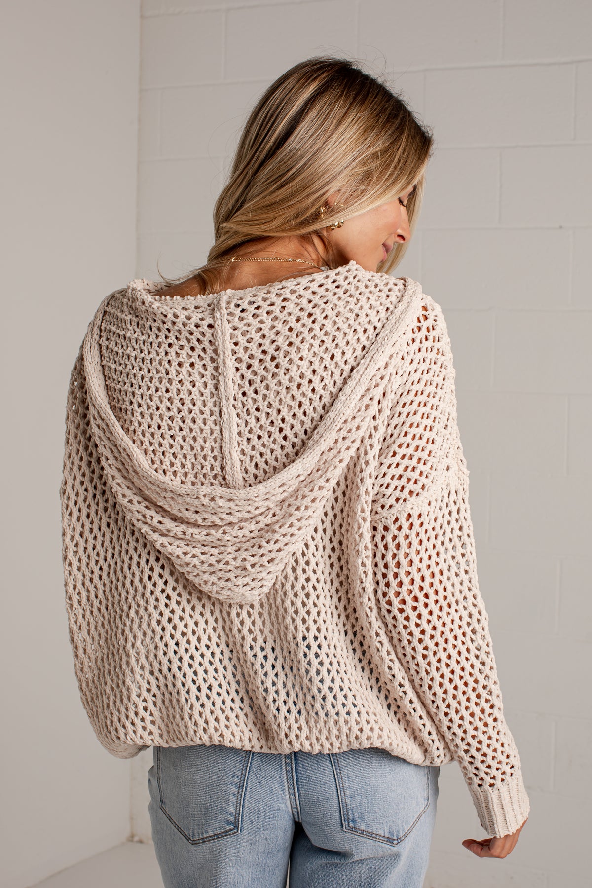 A woman viewed from behind wearing a chunky open-knit hooded sweater in a light beige color, paired with light-wash blue jeans, against a white brick wall.
