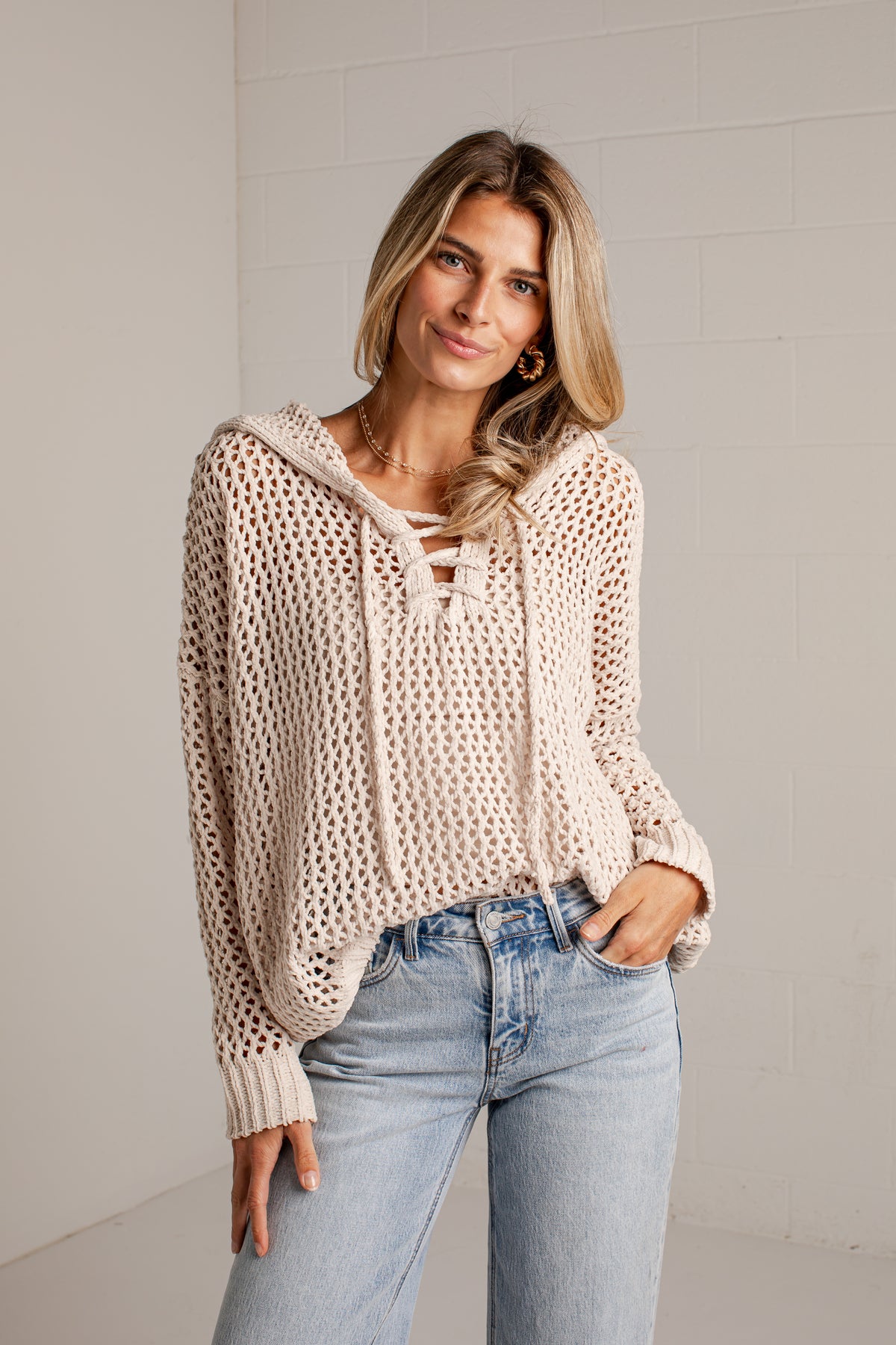 A woman wearing a cream-colored, open-knit hooded sweater with tie-front details and light-wash blue jeans, standing against a light gray brick wall.