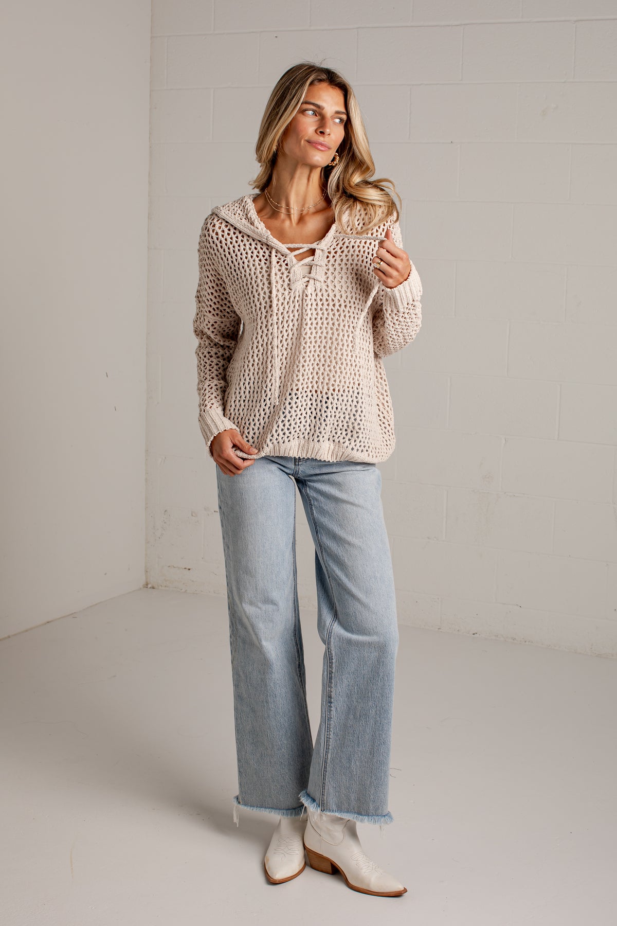 A woman modeling a cream-colored open-knit cropped sweater with a lace-up neckline, paired with light-wash wide-leg jeans and white ankle boots, standing in a minimalist studio setting.