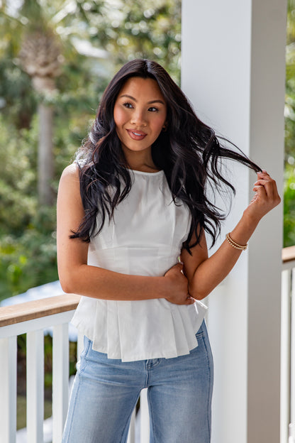 Woman in a white top and blue jeans standing on a porch with greenery in the background