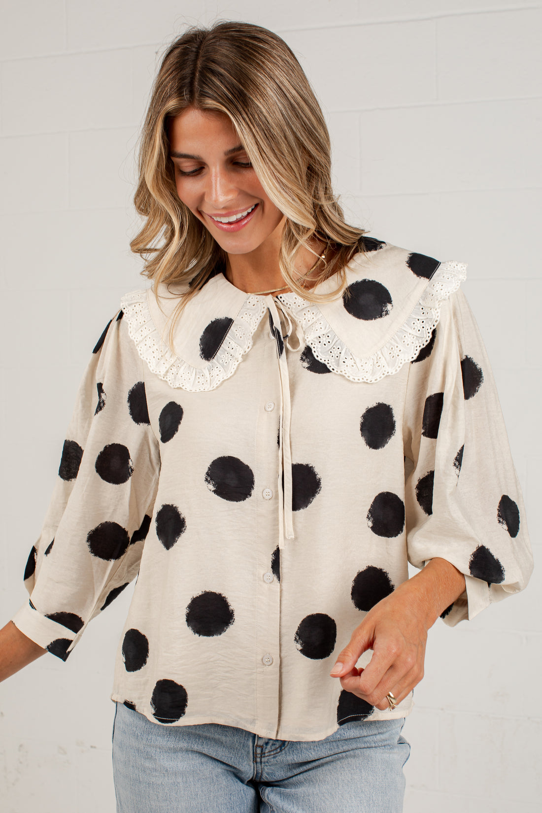 A woman wearing a cream-colored blouse with black polka dots and a white lace Peter Pan collar, paired with light blue jeans, standing against a white brick wall.