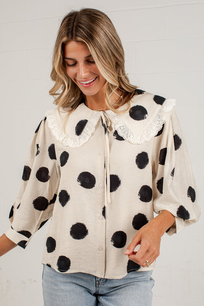 A woman wearing a cream-colored blouse with black polka dots and a white lace Peter Pan collar, paired with light blue jeans, standing against a white brick wall.