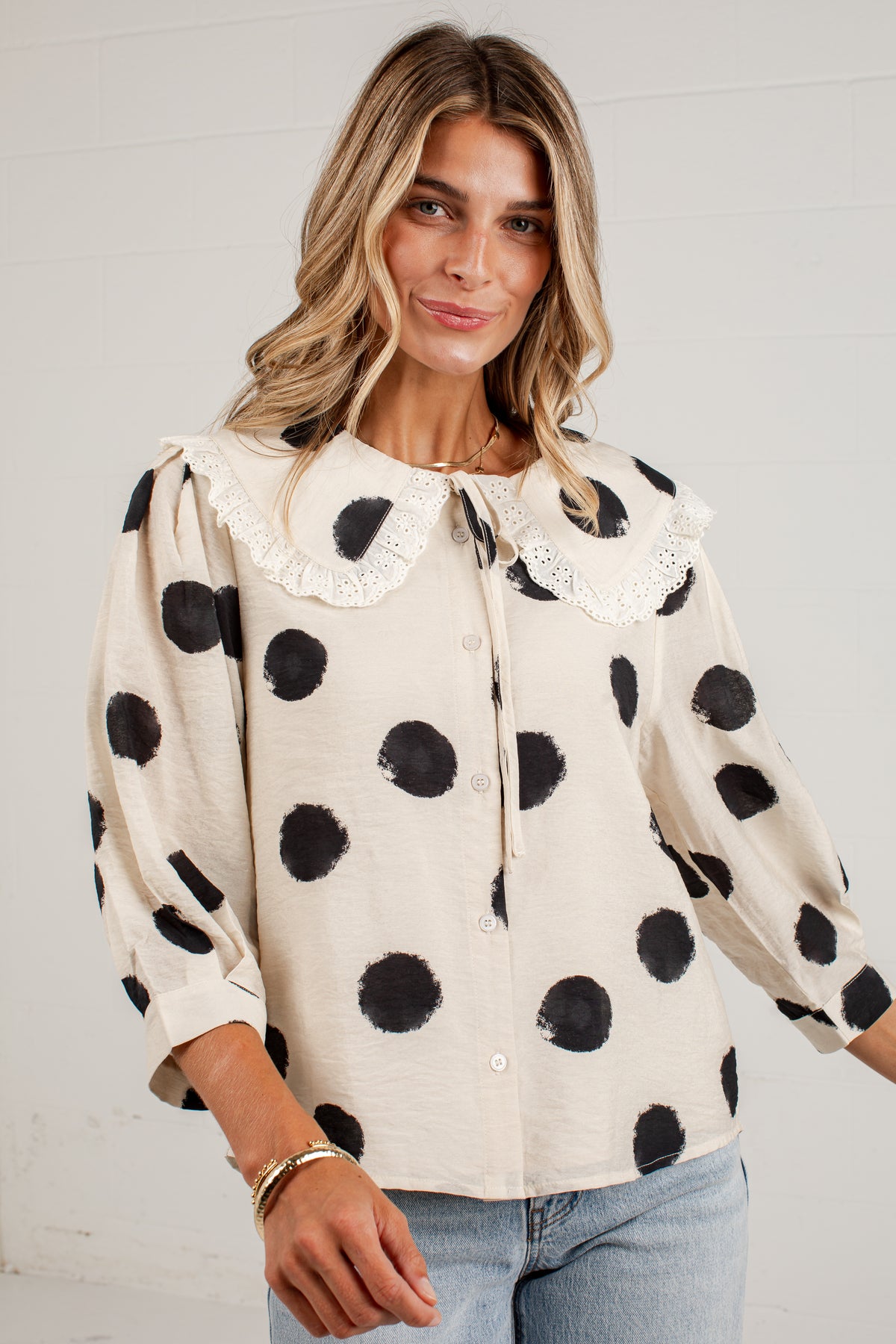 A woman wearing a cream-colored long-sleeve blouse with black polka dots and a large white lace Peter Pan collar, paired with light blue jeans.