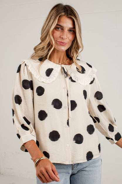 A woman wearing a cream-colored long-sleeve blouse with black polka dots and a large white lace Peter Pan collar, paired with light blue jeans.