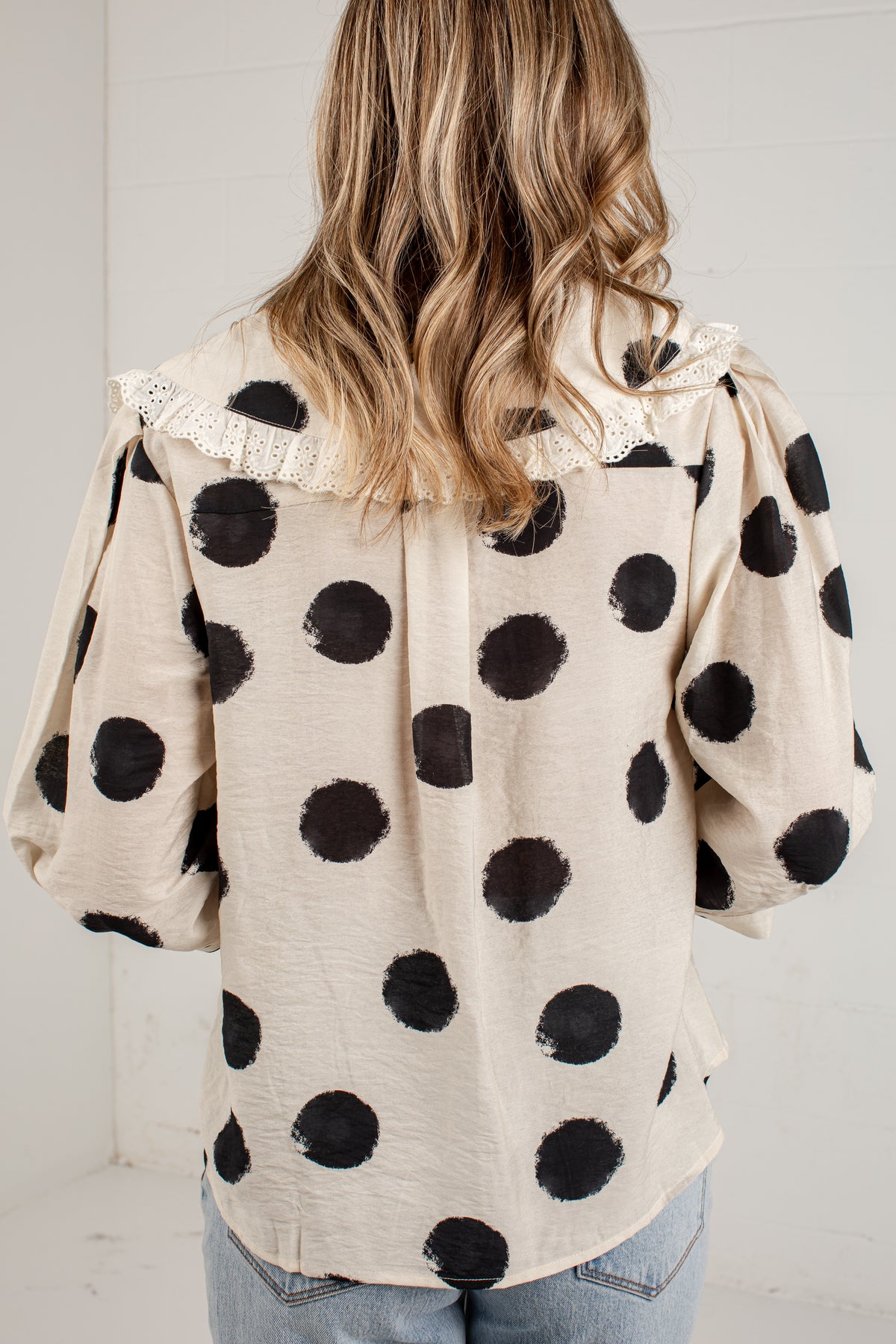 A woman viewed from behind wearing a cream-colored blouse with large black polka dots and a lace collar, paired with light blue jeans.