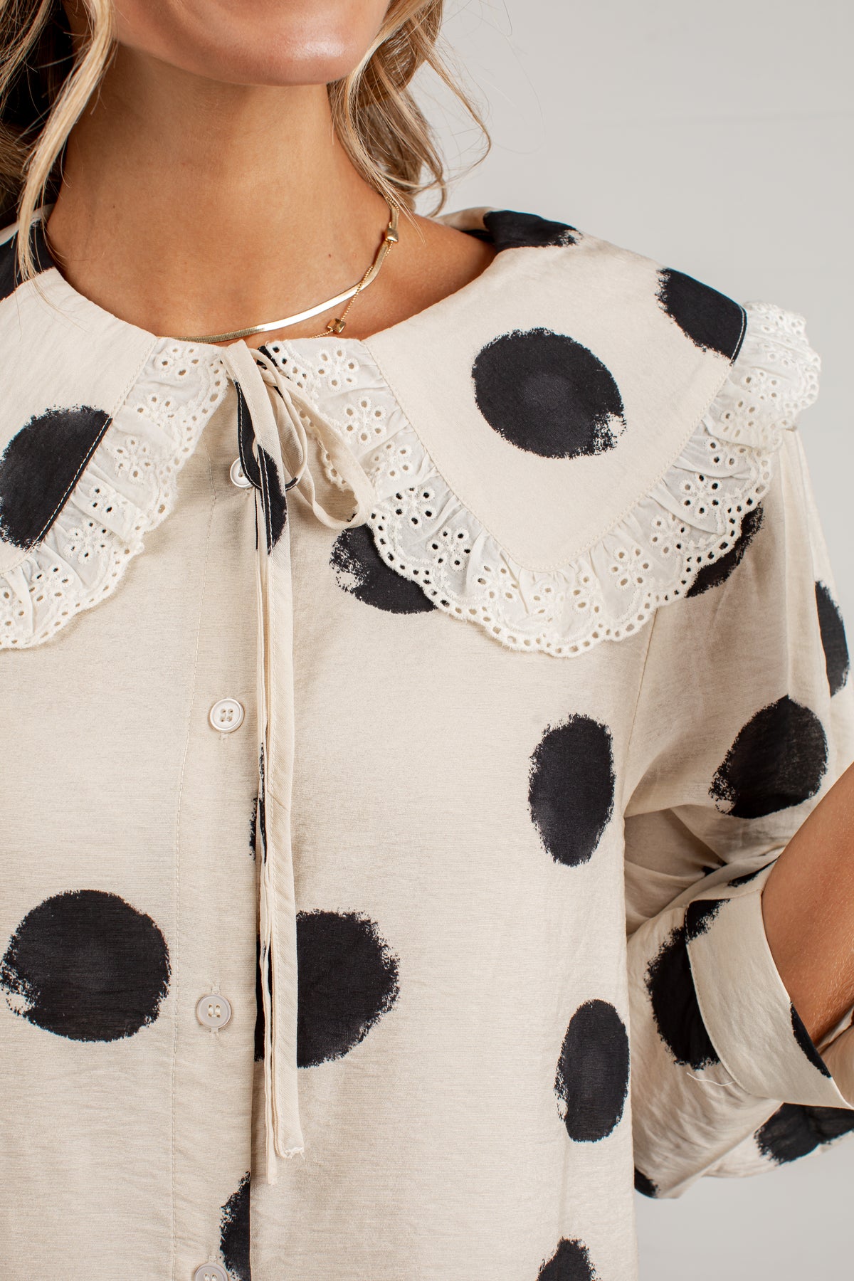 A close-up of a woman wearing a cream-colored blouse with large black polka dots and a wide eyelet lace collar with a tie detail.
