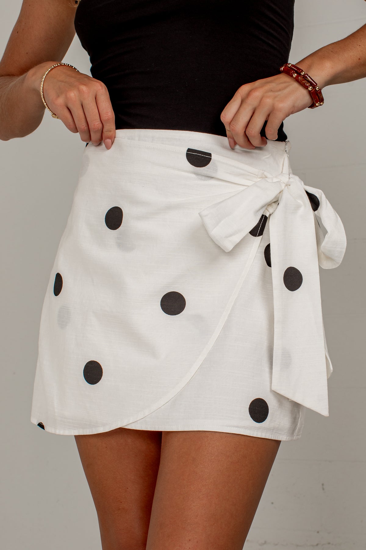 A white wrap-style mini skirt with black polka dots and a tied waist sash, shown on a model wearing a black top against a plain light gray background.