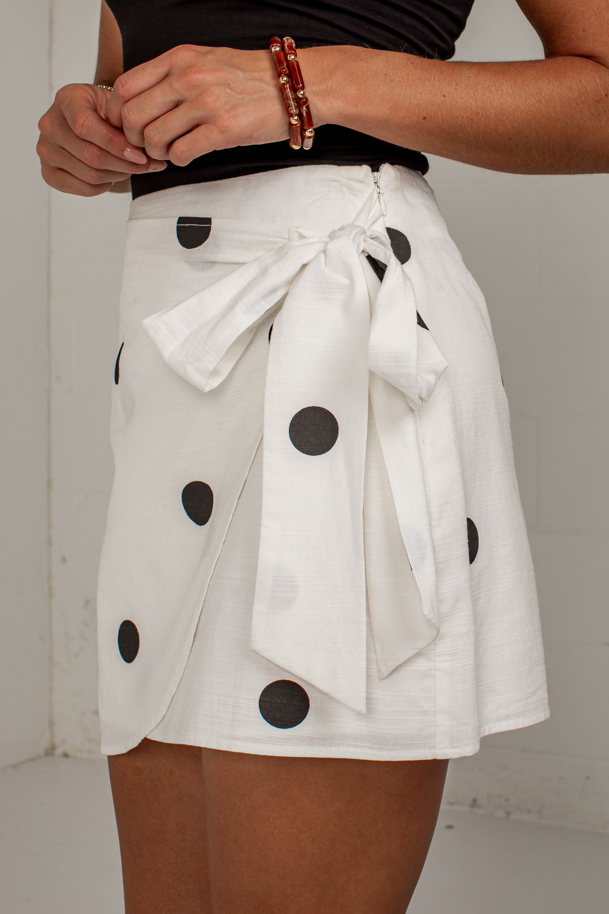 A close-up of a person wearing a white wrap-style mini skirt with large black polka dots and a large tied bow at the front, paired with a black top.