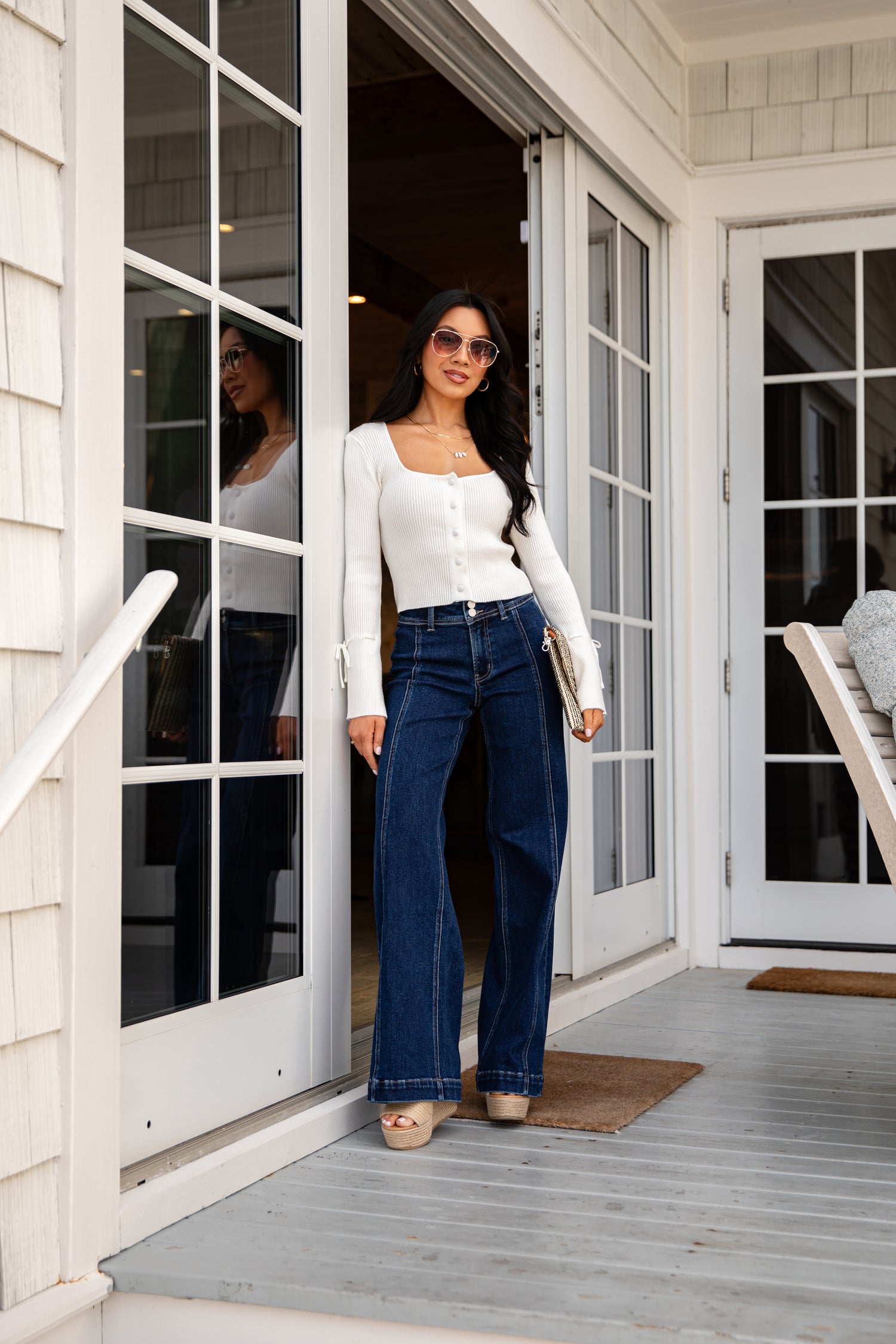 Woman standing on a porch wearing a white top and blue jeans.
